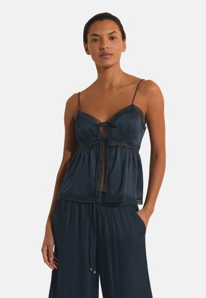 Vrouw draagt een marineblauw mouwloos topje met kanten rand en strikdetail, gecombineerd met bijpassende losse wijde broekspijpen, staand met één hand in de zak.