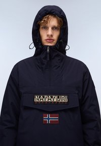 Mørkeblå pullover-jakke med hevet krage, justerbar hette og en lommepose på forsiden. Har brodert logo og Norges flagg.