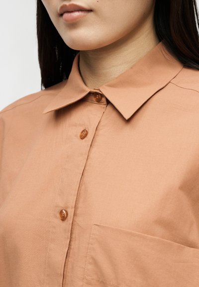Κοντινό πλάνο ενός ατόμου που φοράει ένα ανοιχτό καφέ πουκάμισο με γιακά με μυτερές άκρες και μία μπροστινή τσέπη στο στήθος.