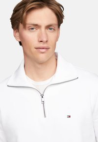Sudadera blanca de media cremallera hecha de una tela suave, con una cremallera plateada y un pequeño logo con acentos en rojo y azul en el pecho.