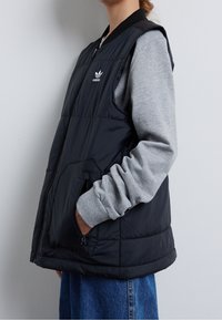 Gilet imbottito nero con zip frontale, tasche laterali e colletto a coste. Indossato sopra una maglietta grigia a maniche lunghe, mostrato su uno sfondo neutro.