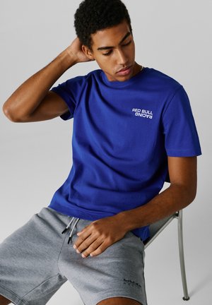 Νεαρός άνδρας καθισμένος σε μεταλλική καρέκλα, φοράει μπλε μπλουζάκι Red Bull Racing και γκρι σορτς, κοιτάζει προς τα κάτω με το ένα χέρι πίσω από το κεφάλι.