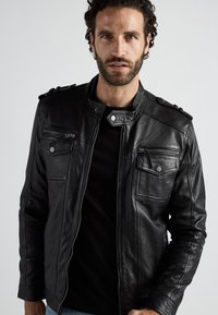 Zwarte leren motorjack met een staande kraag, ritssluiting aan de zijkanten en klepzakken. Heeft schouderepauletten en een gladde textuur.