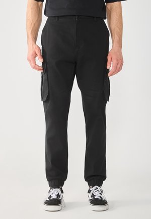 Man draagt zwarte cargobroek met elastische boorden en zwart-witte sneakers, staand tegen een effen lichte achtergrond.