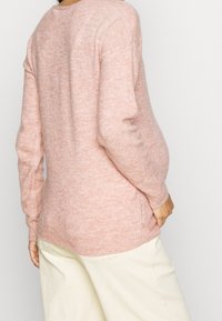 Pull rose tricoté avec une texture douce, col rond, manches longues et ourlet côtelé, associé à un pantalon crème.