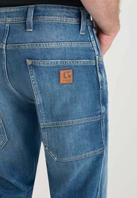 Jeans in denim di un blu medio con una superficie testurizzata. Presentano una tasca con toppa e un'etichetta in pelle con logo sul retro.