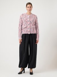 Πλεκτή ροζ και λευκή ζακέτα με υφασματικό μοτίβο, μπροστινά κουμπιά, συνδυασμένη με φαρδιά μαύρη παντελόνια και μαύρες γόβες. Ουδέτερο φόντο.