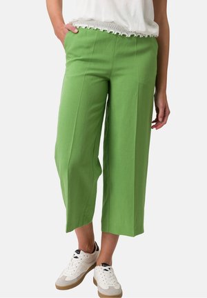 Femme portant un pantalon large court vert, un haut blanc avec un ourlet volanté et des baskets blanches, debout avec une main dans la poche.