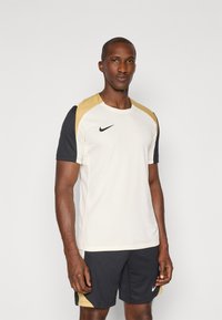 Nike treningsskjorte i krem, med gull- og sorte detaljer. Har korte ermer, rund halslinning og logo på venstre bryst.
