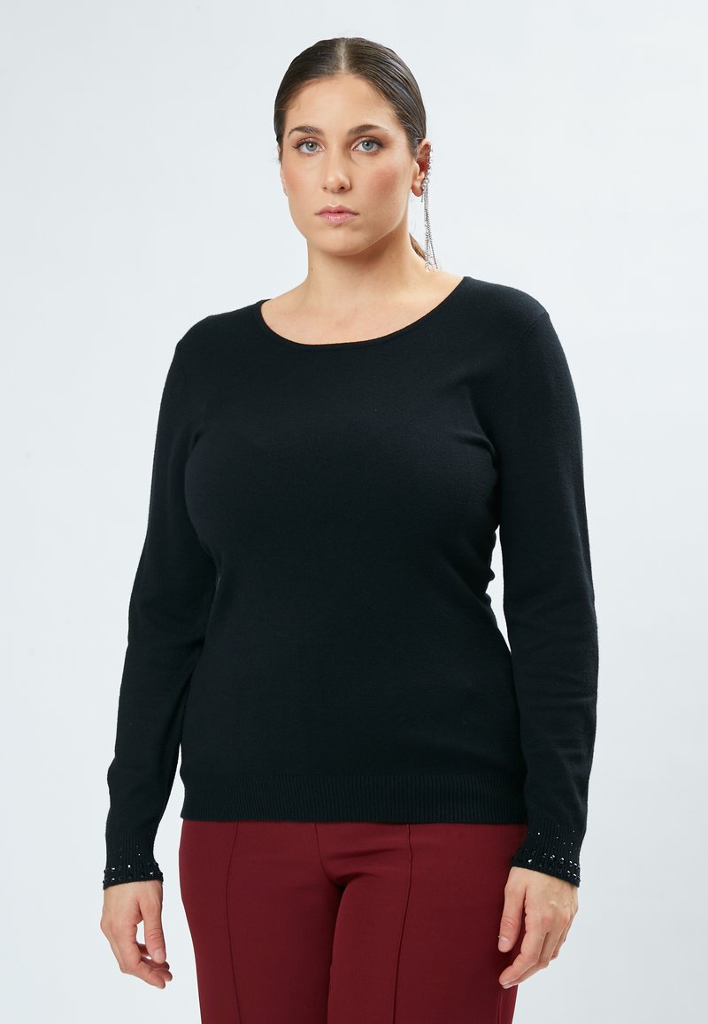 Schwarzer Strickpullover mit langen Ärmeln, Rundhalsausschnitt und strukturierten Bündchen mit kleinen Verzierungen. Kombiniert mit burgunderfarbenen Hosen.