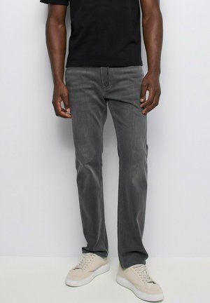 Homme portant un jean droit gris délavé, un t-shirt noir et des baskets écrues, debout contre un mur clair uni.