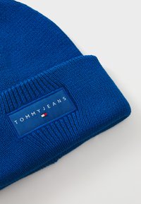 Kék kötött sapka hajtott peremmel, téglalap alakú kék címkével, amelyen a "TOMMY JEANS" felirat található, valamint egy kis piros, fehér és kék logó.