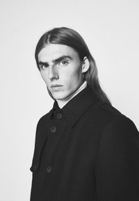 Cappotto di lana nera con chiusura a bottoni, ampio colletto e tessuto testurizzato. Il modello ha i capelli lunghi e indossa un dolcevita chiaro sotto.