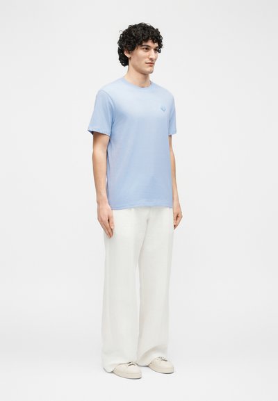 Moški s kodrastimi lasmi, oblečen v svetlo modro srajco s kratkimi rokavi, bele široke hlače in bele superge, stoji ob svetlem enobarvnem ozadju.