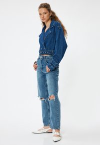 Chaqueta de mezclilla con un diseño corto, color azul, gran cuello, y jeans desgastados con desgarros en las rodillas, combinados con zapatos blancos de punta.