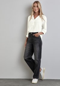 Chemisier blanc à manches longues avec un col à boutons, associé à un jean noir taille haute et des baskets blanches sur un fond gris.