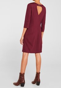 Bordeauxfarbendes Kleid mit Dreiviertelärmeln, ausgestattet mit einem Schlüsselloch-Ausschnitt am Rücken, der durch zwei Knöpfe ergänzt wird. Glattes Gewebe, knielang.
