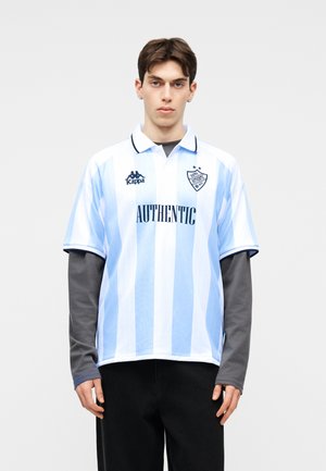 Νεαρός άνδρας που φοράει μπλουζάκι πόλο με κάθετες ρίγες σε ανοιχτό μπλε και λευκό χρώμα, με την επιγραφή "AUTHENTIC", από πάνω ενός μακρυμάνικου γκρι πουλόβερ, σε πόζα.