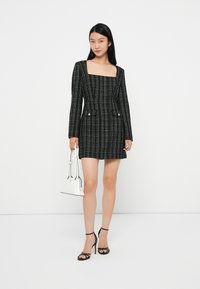 Rochie mini cu carouri alb-negru, cu mâneci lungi, decolteu pătrat și două buzunare frontale. Modelul ține în mână o geantă mică albă și poartă pantofi cu toc negri.