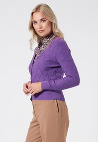 Lila stickad kofta med ett strukturerat diamantmönster, ribbade muddar och fåll. Bärs över en mönstrad blus, tillsammans med beige byxor.