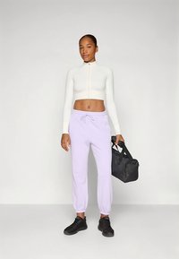 Top bianco a maniche lunghe tagliato, jogging viola chiaro con polsini elasticizzati, scarpe da ginnastica nere, che tiene una borsa da viaggio nera. Design semplice e funzionale.
