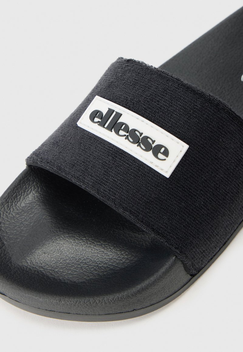 Czarne klapki z teksturowanym paskiem z tkaniny, ozdobionym białą łatą z logo oraz teksturowaną gumową wkładką dla lepszej przyczepności i komfortu.