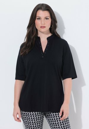 Kvinde med langt mørkt hår iført sort skjorte med knapper og sort-hvide mønstrede bukser, stående foran hvid baggrund.