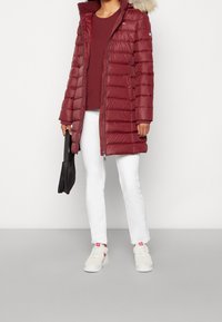 Abrigo puffer rojo con capucha con ribete de piel, con secciones acolchadas, cierre de cremallera y puños de punto. Combinado con pantalones blancos y zapatillas.