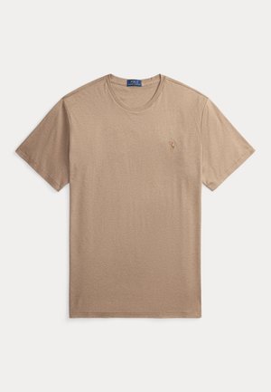Man draagt een beige shirt met korte mouwen, witte broek, bruine riem, bruine schoenen en polshorloge, staat met één hand in zijn zak tegen een neutrale achtergrond.
