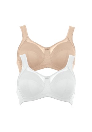 Smiling woman standing barefoot wearing white supportive bra and high-waist underwear against a plain light background.