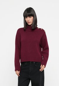 Vinröd tröja med hög hals, ribbad textur och guldknappar på axlarna. Kombineras med svarta jeans.