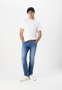 Camiseta blanca de manga corta con pequeño logo, jeans azul claro, cinturón marrón y zapatillas blancas. Atuendo casual simple con un corte relajado.