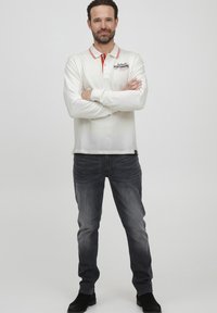Langærmet hvid polo med røde kraveaccenter, prydet med tekstdetaljer; parret med mørkegrå jeans og sorte støvler. Enkel, afslappet design.