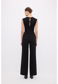 Combinaison noire avec un design sans manches, jambes larges et un détail de dos en forme de trou de serrure. Tissu lisse avec une taille cintrée et un style minimaliste.