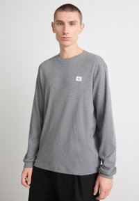 Γκρι μακρυμάνικο T-shirt φτιαγμένο από υφαντό ύφασμα με στρογγυλή λαιμόκοψη και μια μικρή πατίνα με λογότυπο στο αριστερό στήθος.