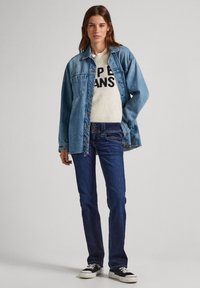 Mulher em pé, vestindo uma jaqueta de ganga azul sobre um suéter branco com texto preto, jeans azul-escuro e ténis em preto e branco.