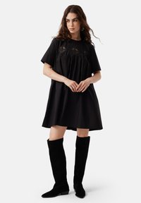 Rochie din bumbac negru cu mâneci scurte, design plisat și detalii complexe din dantelă pe bust, purtată cu botine negre din suède până la genunchi.