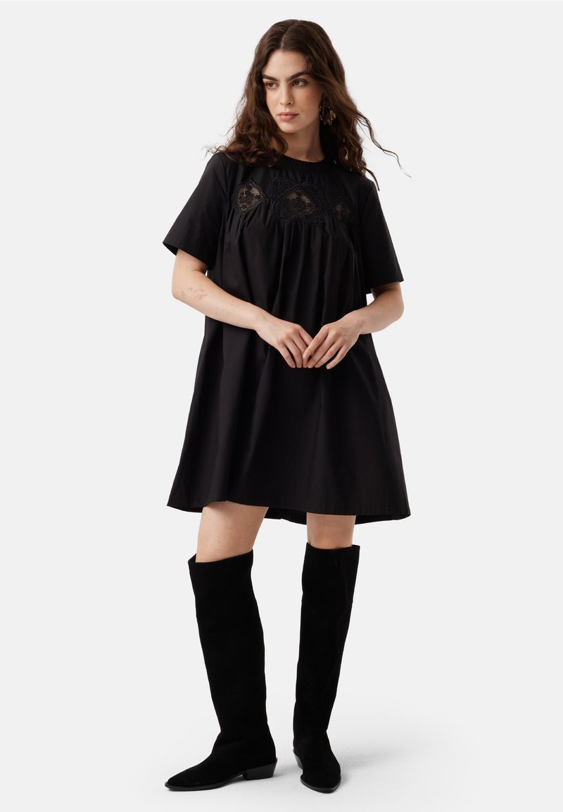 Rochie din bumbac negru cu mâneci scurte, design plisat și detalii complexe din dantelă pe bust, purtată cu botine negre din suède p�ână la genunchi.