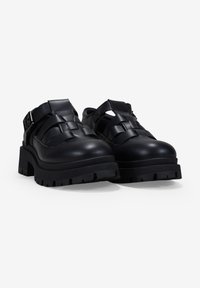 Zapatos de cuero negro con un diseño troquelado, suela gruesa y texturizada, y detalles de hebilla, combinando estilo con una apariencia robusta.