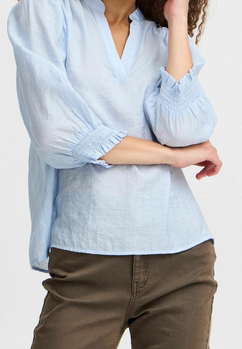 Blouse bleu clair, ample, avec un décolleté en V et des manches bouffantes, présentant des accents froncés aux poignets et un tissu texturé.