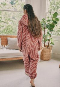 Fleece onesie in pink with a zebra pattern, featuring a hood, long sleeves, and elastic cuffs. The fabric is soft and textured.