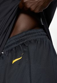 Juodi sportiniai šortai su tekstūruotu audiniu, su elastine juosta ir mažuoju geltonu Nike swoosh logotipu apatinėje pusėje.