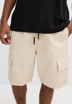 Hombre vistiendo pantalones cortos cargo beige con bolsillos laterales y camiseta negra, manos descansando cerca de los bolsillos sobre un fondo liso.