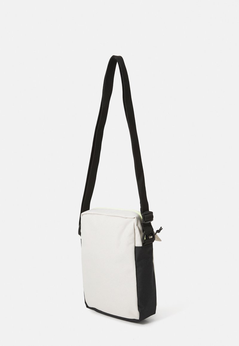 Bolso cruzado rectangular pequeño con frente beige y lados y asa negros, mostrado colgado sobre un fondo blanco liso.