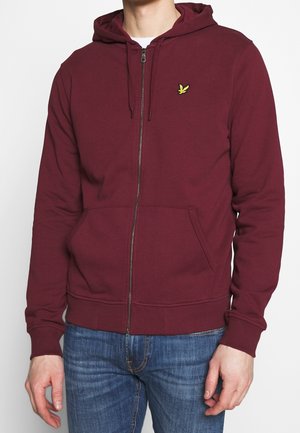 Mann trägt einen weinroten Hoodie mit Reißverschluss und kleinem gelbem Vogel-Logo auf der Brust sowie blaue Jeans, steht mit entspannt an den Seiten hängenden Händen.