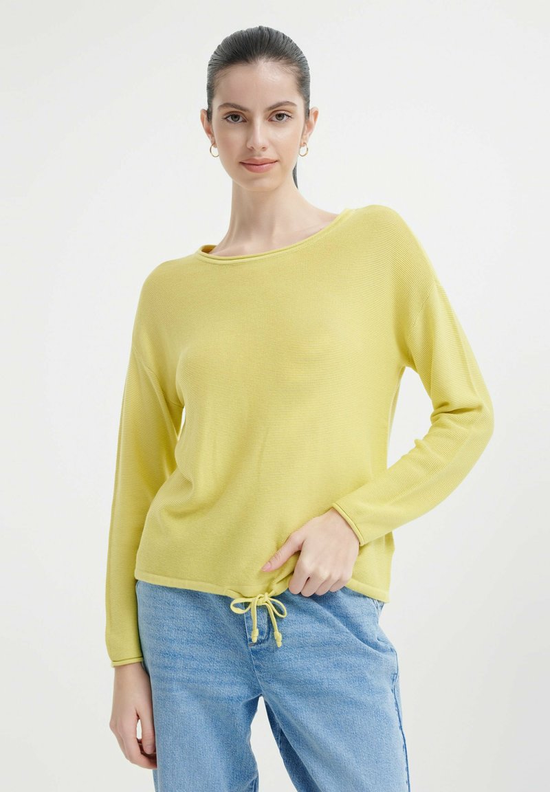 Geel lang mouwtop met een ronde hals en gestructureerde stof, met een trekkoord aan de onderkant en een ontspannen pasvorm, gecombineerd met blauwe jeans.