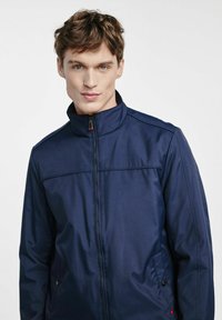 Granatowa kurtka zapinana na zamek z wysokim kołnierzem, gładką fakturą, kieszeniami z przodu i czerwonym akcentem na uchwycie zamka. Prosty, schludny design.