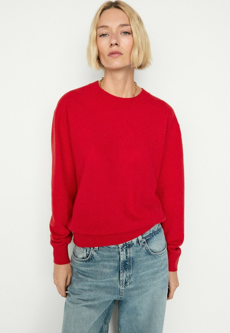 Personne aux cheveux blonds courts, portant un pull rouge vif et un jean bleu clair, debout devant un fond blanc uni.