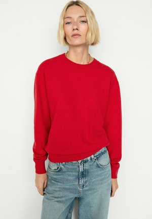Person med kort blond hår iført en klar rød sweater og lyseblå jeans, stående mod en helt hvid baggrund.