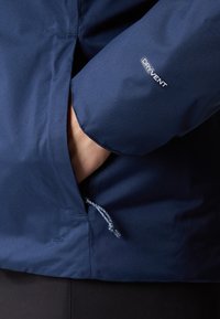 Jūrinis vandeniui atsparus džinsas, pasižymintis tekstūruotu audiniu, reguliuojamu raišteliu apatine dalimi ir "DRYVENT" logotipu ant rankovės.